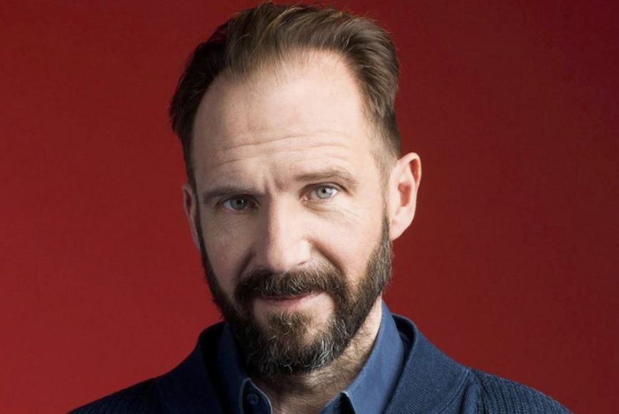 Actor Ralph Fiennes smiling at the camera in front of a red background